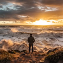 Raging ocean with big waves, a man in a sand-colored jacket stands facing the ocean, autumn weather, sunset light