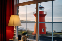 The warm interior of a room with a view of a striking red lighthouse standing guard by the sea at dusk, a scene of serene maritime navigation.