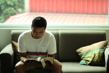 a man reading a Bible sitting on a couch in his living room 