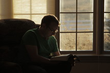 Man sitting in a chair in front of a window reading the Bible.