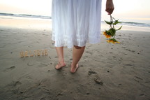 word BELIEVE on sand and woman in bare feet holding flowers