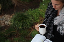 a woman standing outdoors in a scarf holding a mug and reading a Bible 