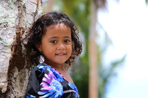 A young polynesian girl with curly hair, leaning against a tree, dressed for Church 