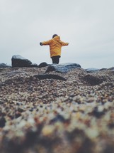 child in a raincoat standing on a shore with outstretched arms 