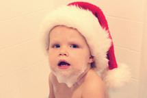 A little boy with soap suds on his chin and wearing a Santa hat.