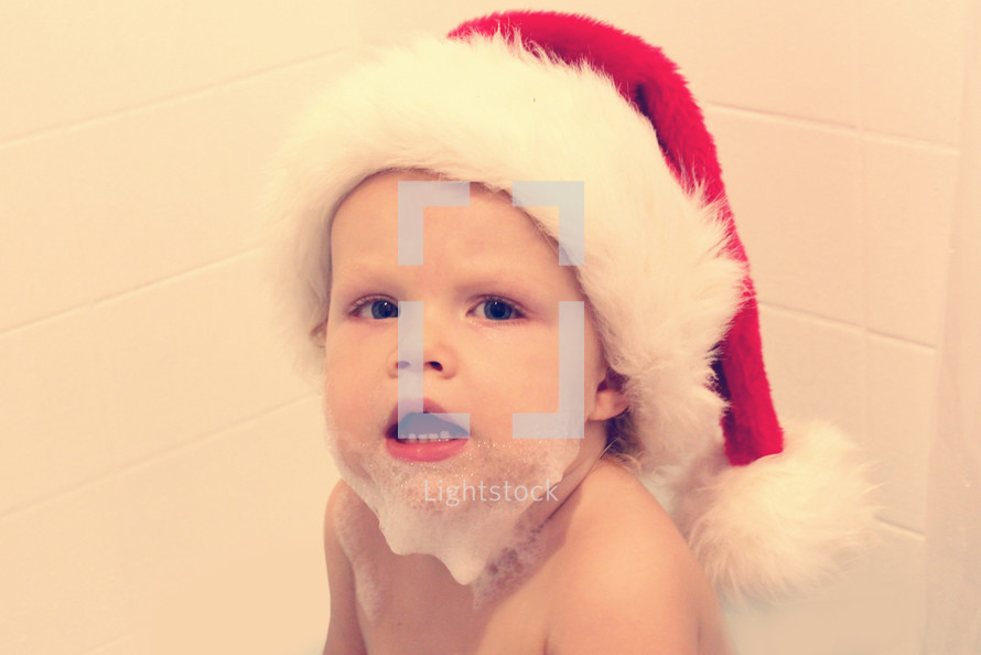 A little boy with soap suds on his chin and wearing a Santa hat.