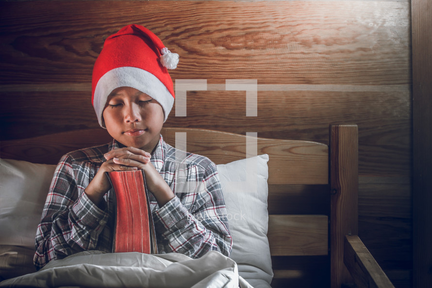 a boy in a Santa hat reading a Bible and praying in bed 