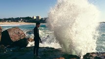 a man in a wetsuit standing at the edge of a rocking shore with crashing waves