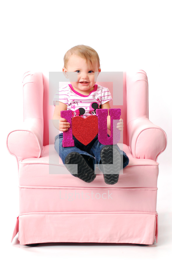 toddler sitting on a pink recliner holding an I heart you sign 
