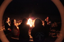 halo around a group of friends sitting around a campfire on a beach at night 