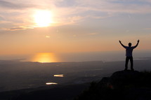 silhouette of a man standing on a mountain top with raised hands in surrender to God