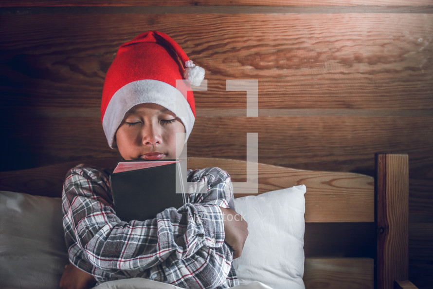 a boy in a Santa hat hugging a Bible 