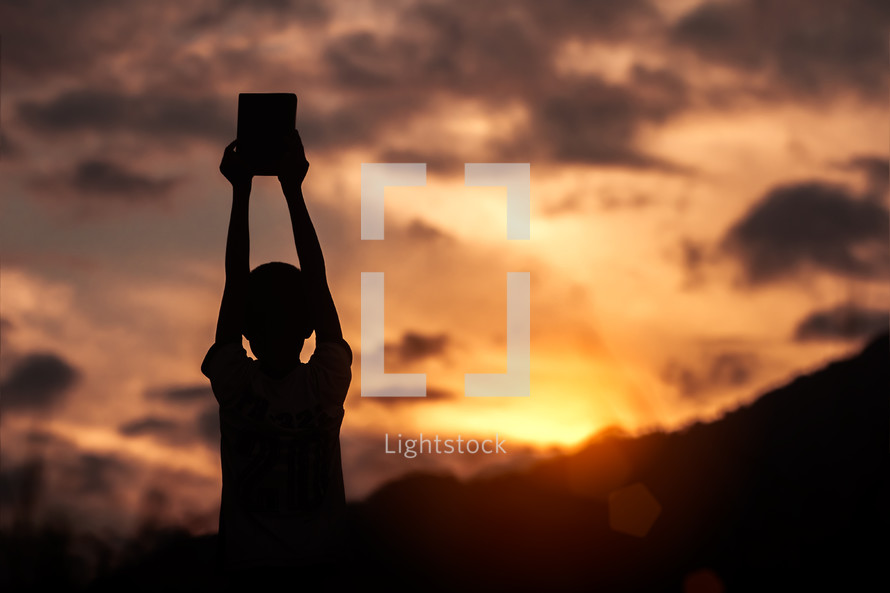 silhouette of a boy holding up a Bible at sunset 
