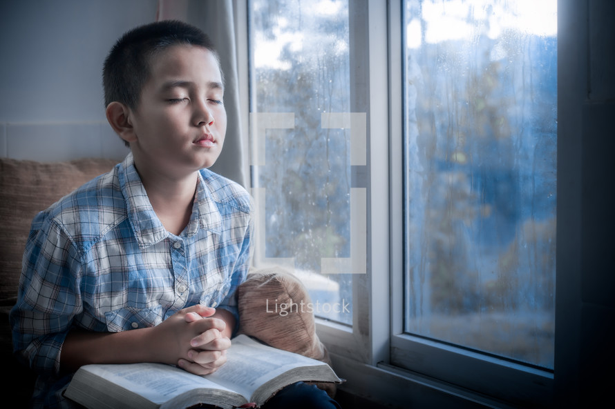 a boy sitting in a window praying over a Bible 