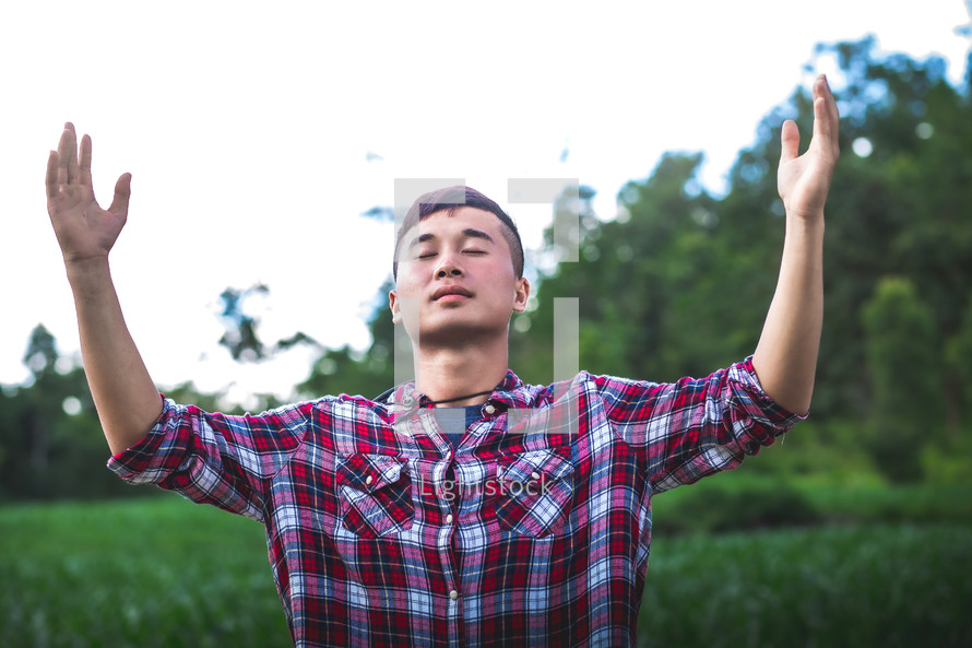 young man in a plaid shirt standing outdoors with hands raised 
