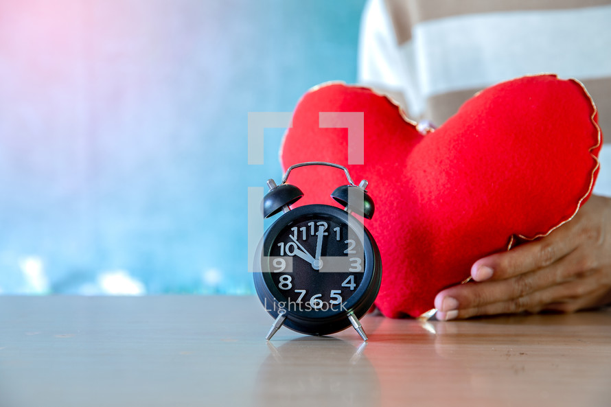Alarm clock on wooden table with a red heart in hands, valentine day background.