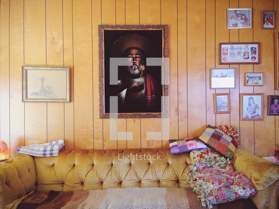 Painting of Christ hanging on a paneled wall, surrounded by family photos, in a den with a velvet sofa covered in quilts.