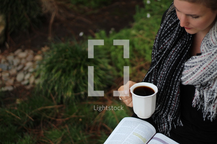 a woman standing outdoors in a scarf holding a mug and reading a Bible 