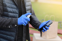 Delivery service courier during the Coronavirus, COVID-19, pandemic, cropped courier hands in gloves spraying alcohol disinfectant spray on cellphone near cardboard boxes outdoors
