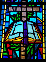 A colorful stained glass window depicting the Cross and the word of God in vivid blue, gold, green and red colors adorning a church sanctuary. 