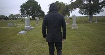 Sombre, sad young man in black suit walking in cinematic slow motion in cemetery through graveyard tombstones.