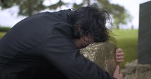 Sad, grieving man embracing family tombstone in cemetery crying in cinematic slow motion. 