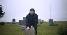 Young, sad man in black suit kneeling in cemetery at graveyard tombstone grieving in cinematic slow motion.