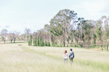 Couple walking in a grassy field near a tree line.