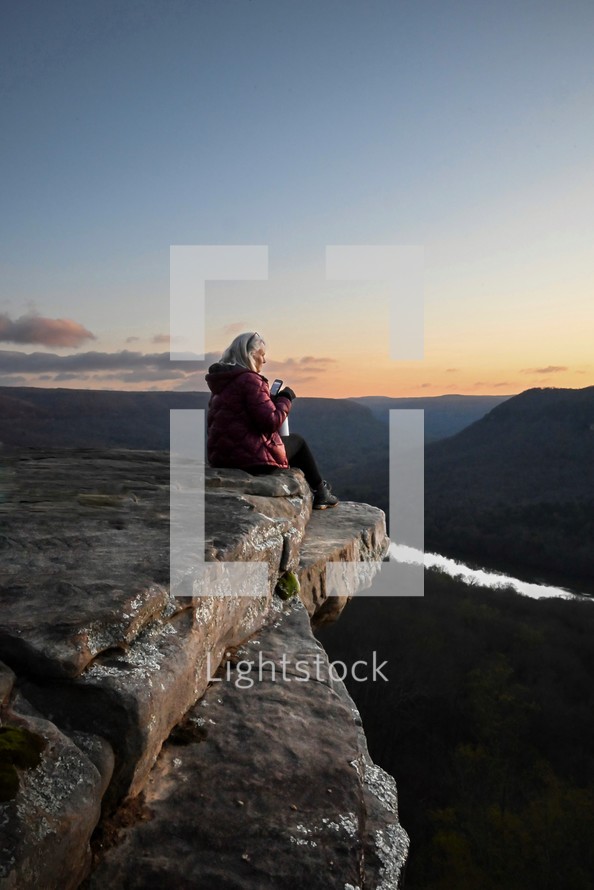 a woman sitting at the edge of a cliff at sunrise 