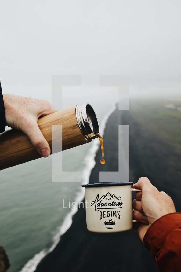 Two hands pouring coffee into the adventure begins mug above a black sand beach in Iceland