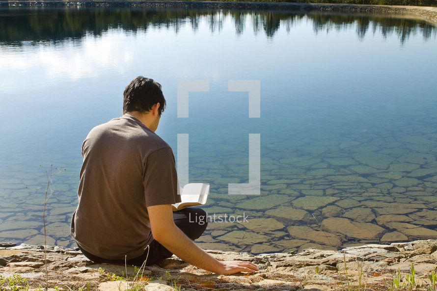 man reading a Bible sitting by a lake