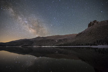 The milky way stretches is reflected in a crystal-clear lake under the watch of a rocky peak covered in snow.  