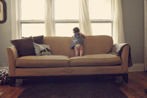 Toddler child standing on a sofa looking out the window.