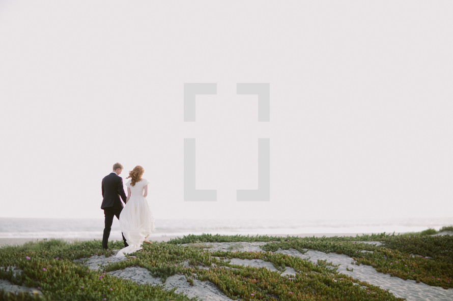 a bride and groom walking  towards a beach 