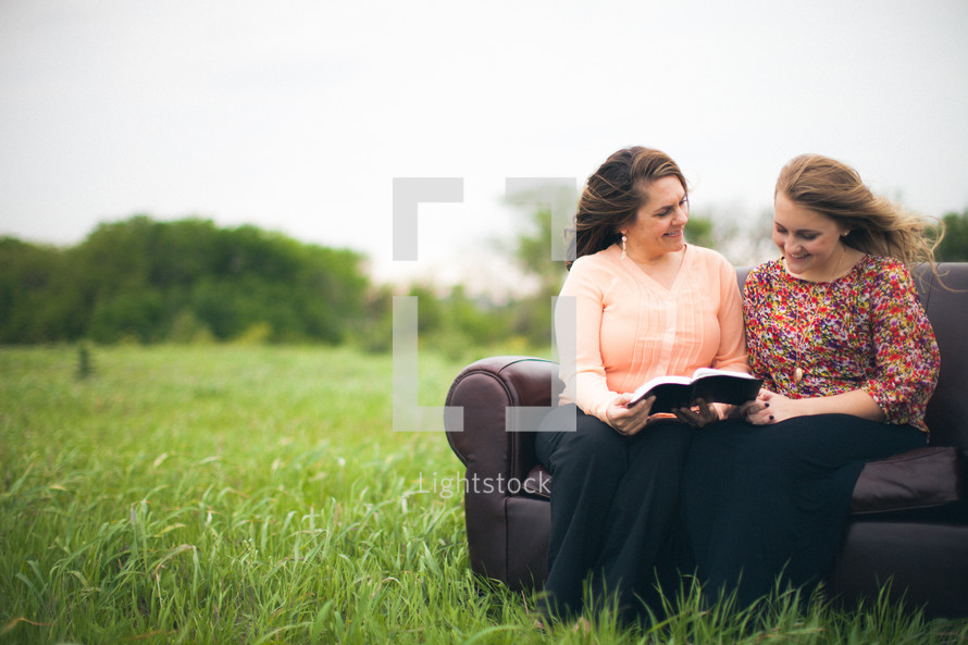 Smiling mother and daughter reading the Bible together while sitting on a sofa in a field.