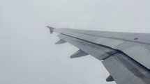 The wing of a flying passenger aircraft with closed flaps against a backdrop of gray sky and dense clouds