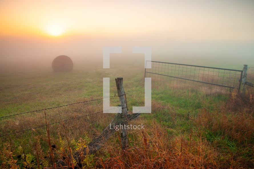 Open gate entrance into grassy field with hay bale at sunrise with morning ground fog