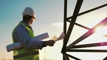 Foreman stands on construction site and holds drawings on paper in hands. Man shows hand where future construction be according to plan. Field with telecommunications tower. Male in protective helmet