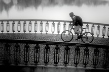 Unidentifiable hooded cyclist crossing a paved old bridge. Black and white image
