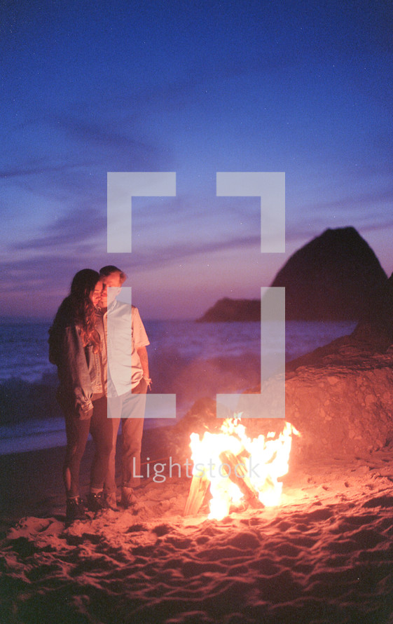 a couple standing by a fire on a beach 