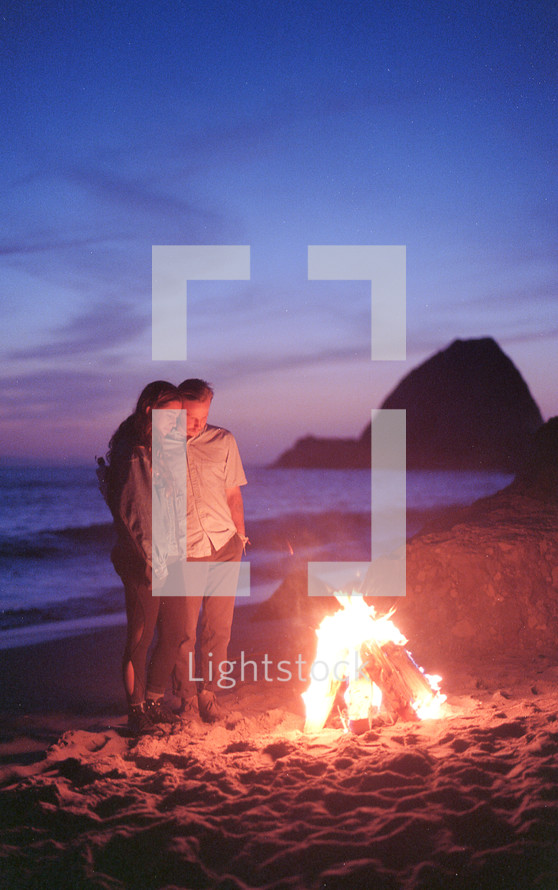 a couple standing by a fire on a beach 