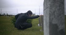 Young, sad man in black suit kneeling in cemetery at graveyard tombstone grieving in cinematic slow motion.