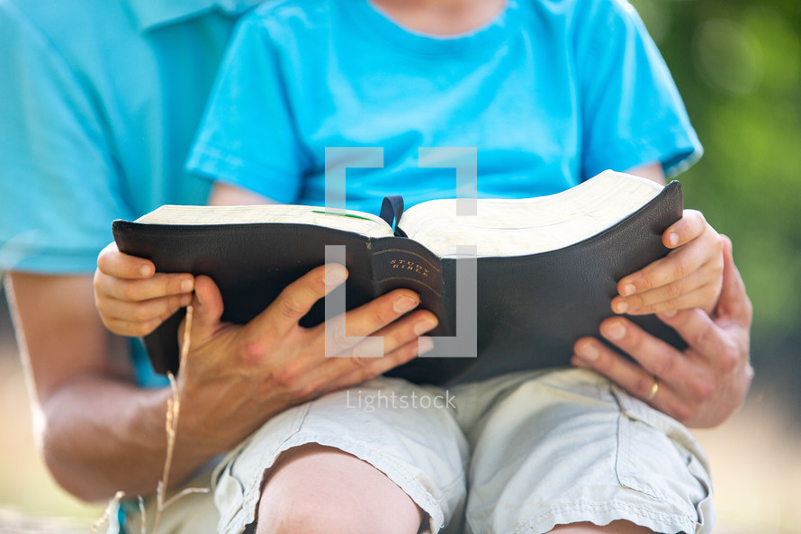 Father and young son on his lap reading the Bible together outside