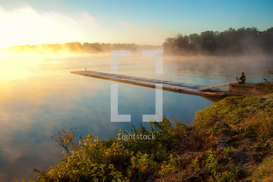 Man on wooden boardwalk pier photographing peaceful foggy lake at sunrise