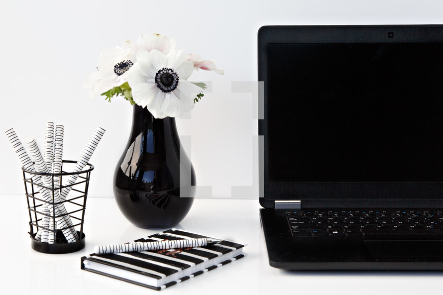 flowers in a black vase, journal, pens, and laptop computer on a desk 