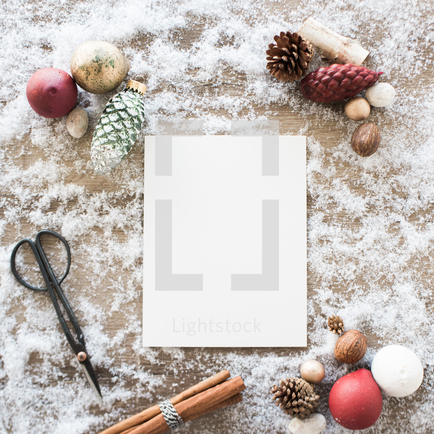 white paper in snow with border of scissors, cinnamon sticks, and pine cones 