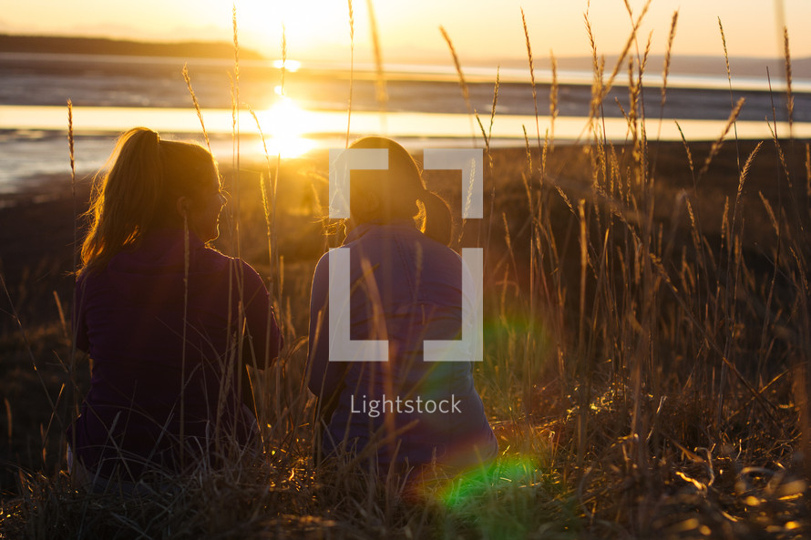 Two girls sit in tall grass facing a glowing sunset.