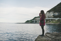 a woman standing on a rock by a seaside highway 