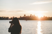 woman standing across the river from a city taking a picture 