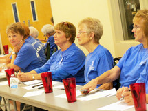 A group of ladies greet guests at a church sponsored women's retreat at a registration volunteer desk wearing blue shirts to help sponsor and advertise the event.  